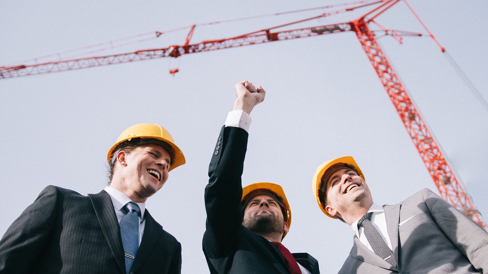 Three businessmen laughing in front of a crane. One has their hand raised in a fist.