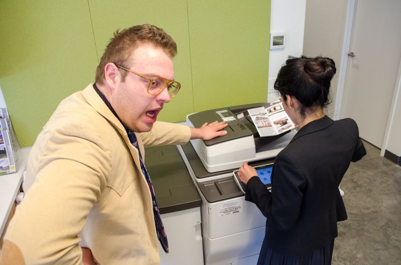 Two employees standing by a fax machine, one is upset that they are having to wait for the other.