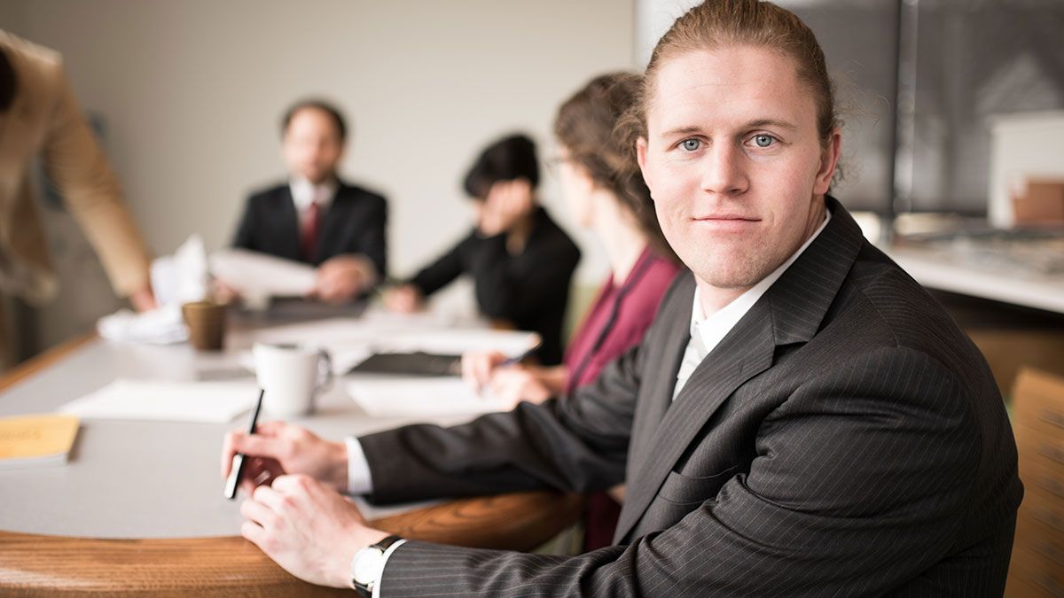 A businessman smiling at the camera while others work in the background in a board room.