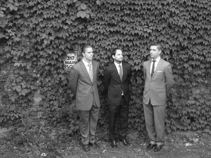 3 businessmen standing outside by leaves on a brick wall